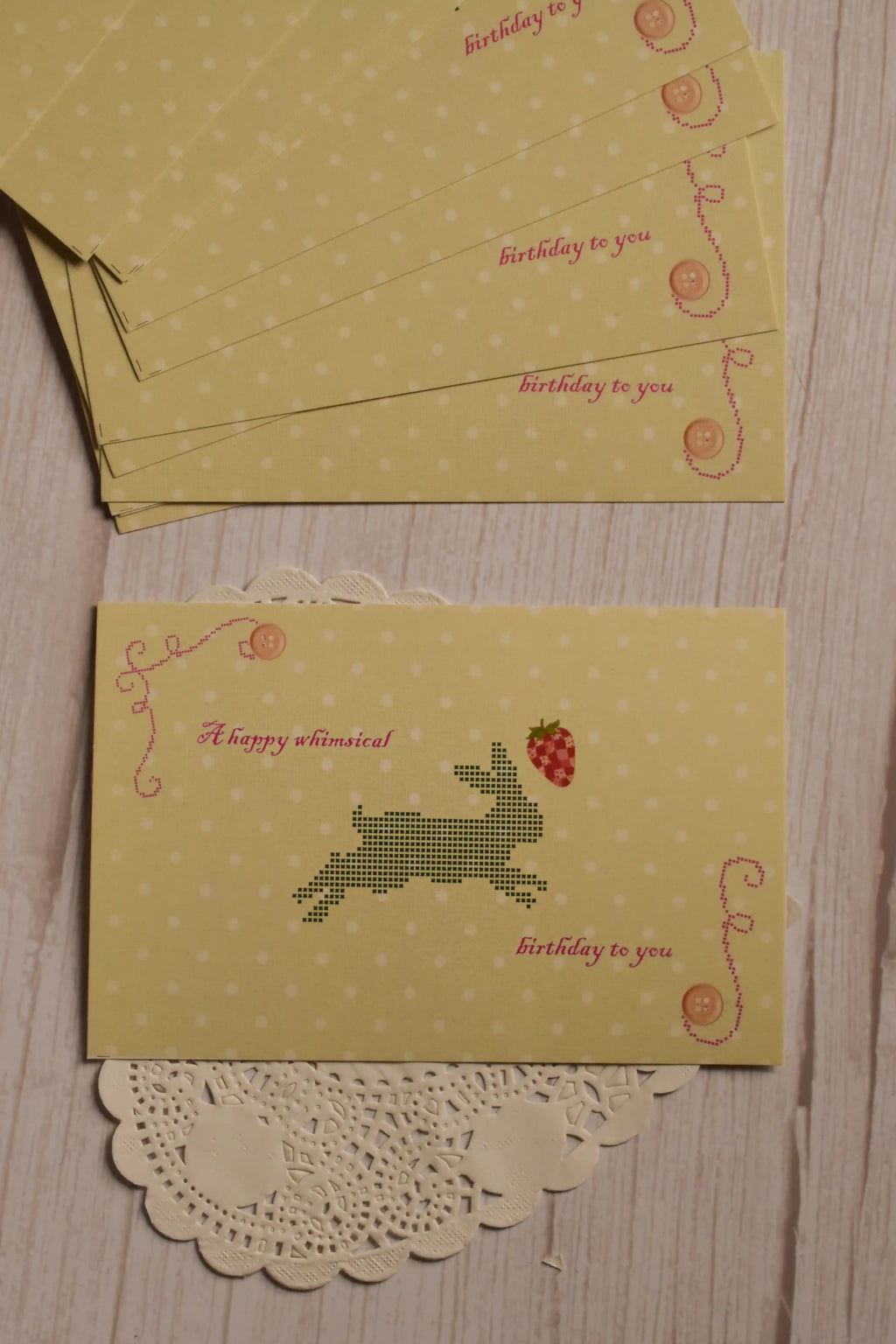 The front of the yellow birthday postcard displayed with multiple matching postcards, featuring a pixel-style rabbit, strawberry, button details and the words “A happy whimsical birthday to you.”