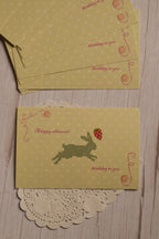 The front of the yellow birthday postcard displayed with multiple matching postcards, featuring a pixel-style rabbit, strawberry, button details and the words “A happy whimsical birthday to you.”