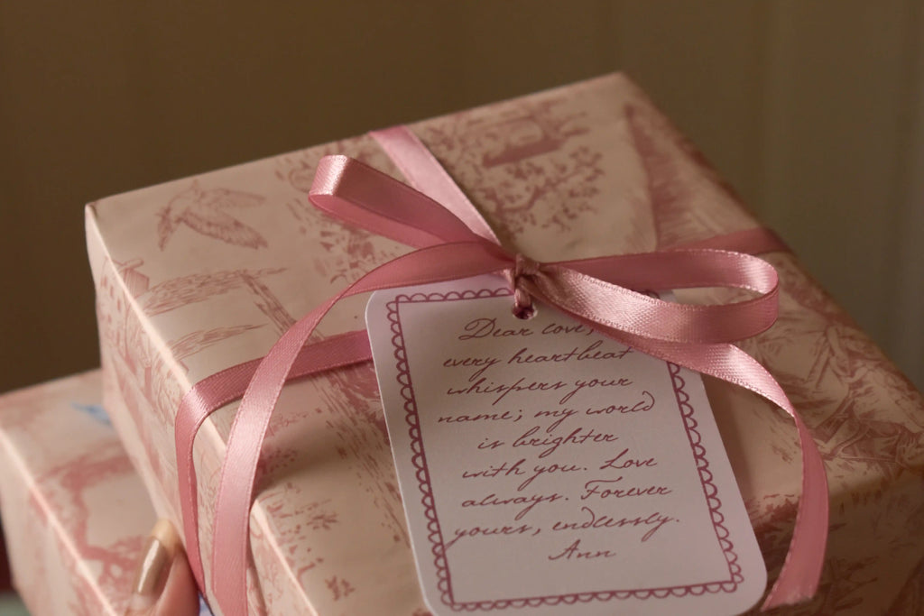 A softly lit gift presentation featuring vintage toile wrapping paper, a rose pink ribbon tied into a bow and a decorative message tag in coordinating pink tones.