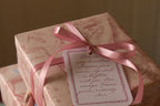 A softly lit gift presentation featuring vintage toile wrapping paper, a rose pink ribbon tied into a bow and a decorative message tag in coordinating pink tones.