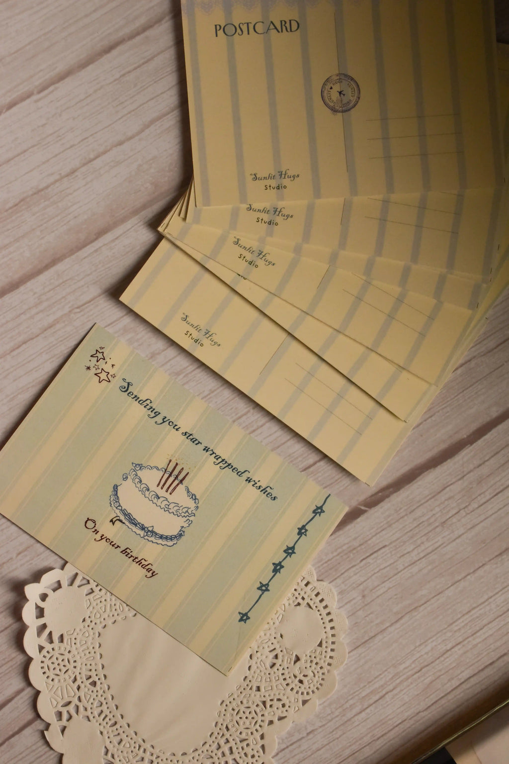 A styled arrangement showing both sides of the Star-Wrapped Wishes Birthday Postcard. The front features a cake and celestial birthday message, while the back includes a classic striped postcard layout.