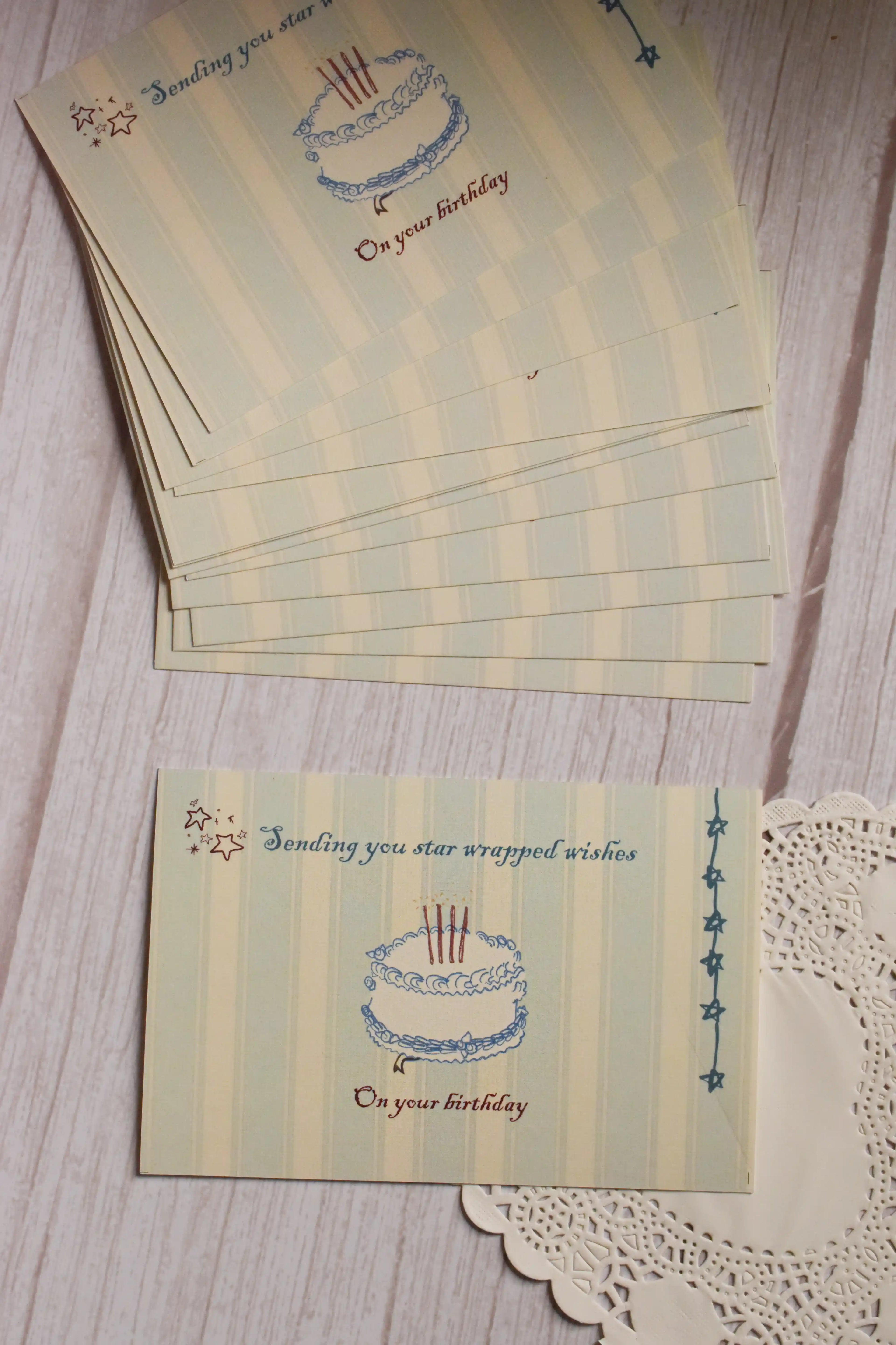 Front view of the Star-Wrapped Wishes Birthday Postcard displayed beside a stack of matching postcards. The vintage-inspired design features an illustrated birthday cake, tiny stars and the message “Sending you star wrapped wishes on your birthday.”