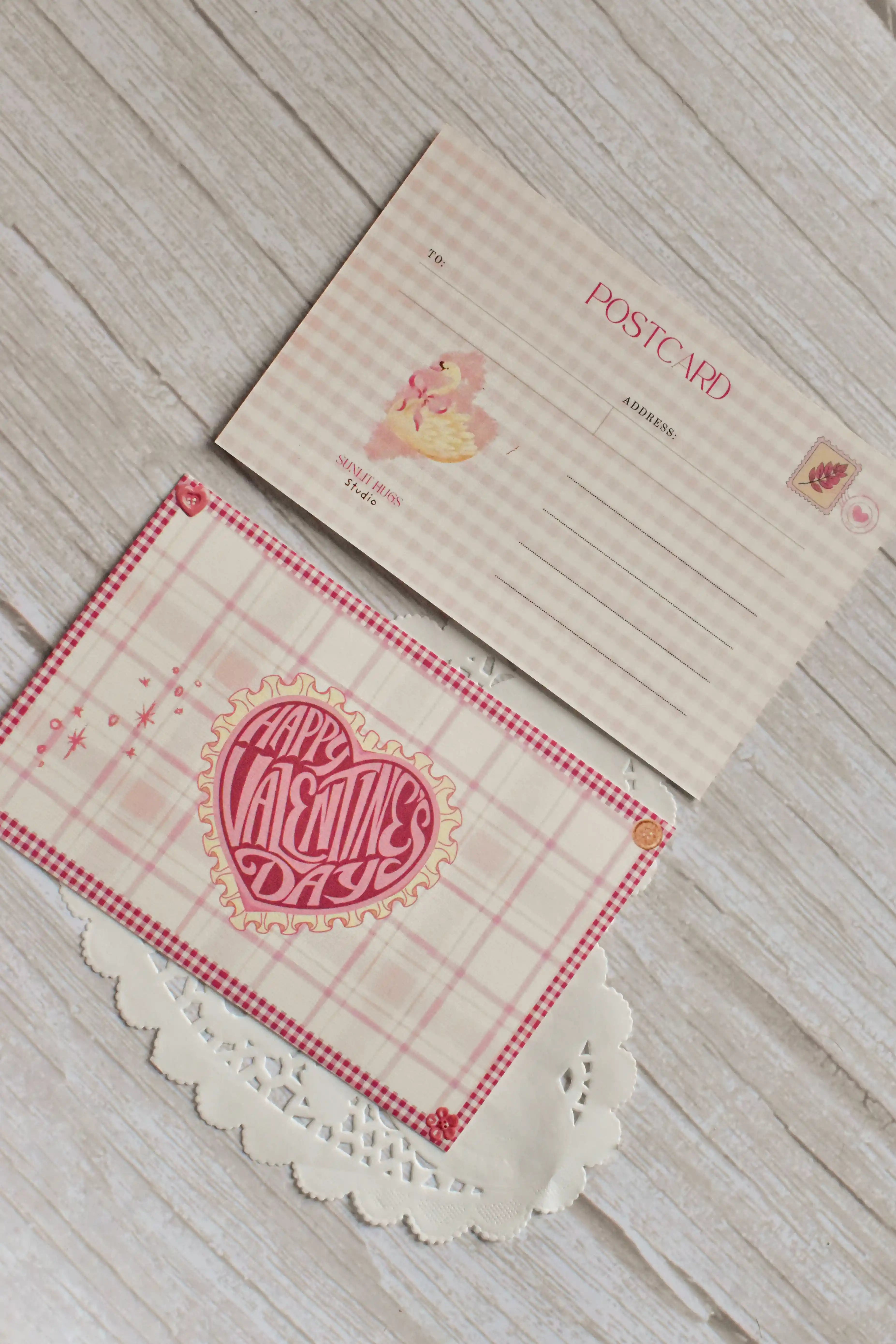 A pink gingham Valentine postcard shows vintage heart and swan artwork on a tabletop.