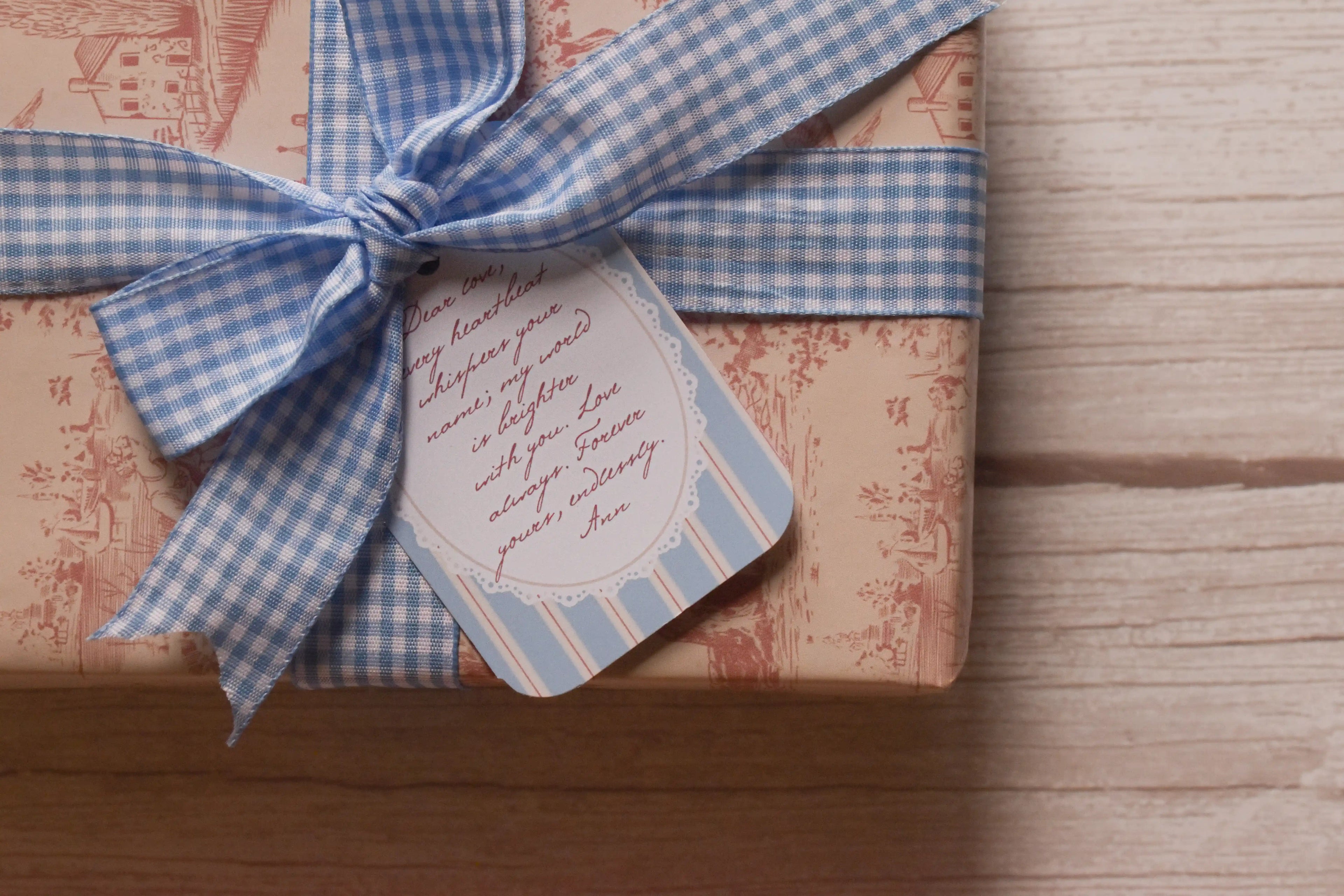 Close-up of soft cream and muted rose vintage toile gift wrapping, finished with a powder blue and white gingham ribbon bow and a coordinating blue striped message tag.