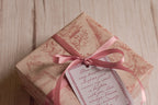 A gift wrapped in soft cream and dusty rose toile-style paper featuring birds, trees, figures and vintage architecture, finished with a rose pink ribbon bow and decorative message tag.