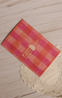 Angled close-up of the postcard front featuring a hanging jellyfish, tiny celestial details and the phrase “You fill this world with so much magic” over a coral and pink checkered design.