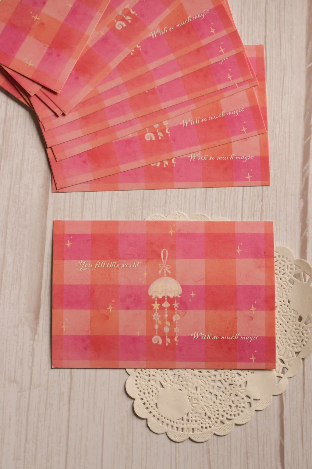 Front view of the 4 x 6 inch postcard featuring a pink and coral checkered background, delicate stars, a hanging jellyfish illustration and the words “You fill this world with so much magic.”