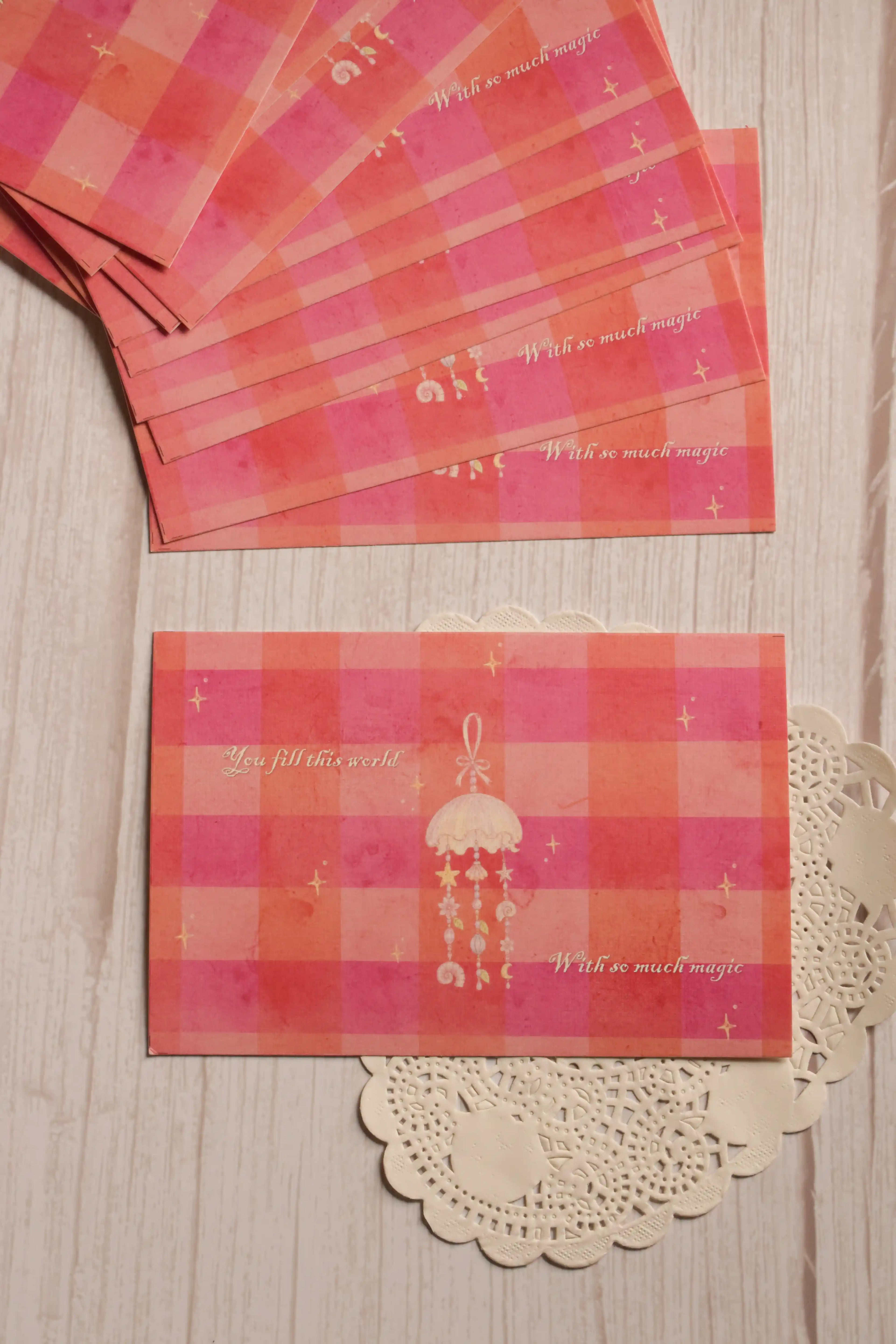 Front view of the 4 x 6 inch postcard featuring a pink and coral checkered background, delicate stars, a hanging jellyfish illustration and the words “You fill this world with so much magic.”