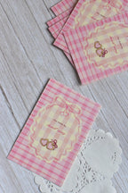A Valentine postcard with pink gingham, red cherries, and bow accents is shown in a romantic flat lay.