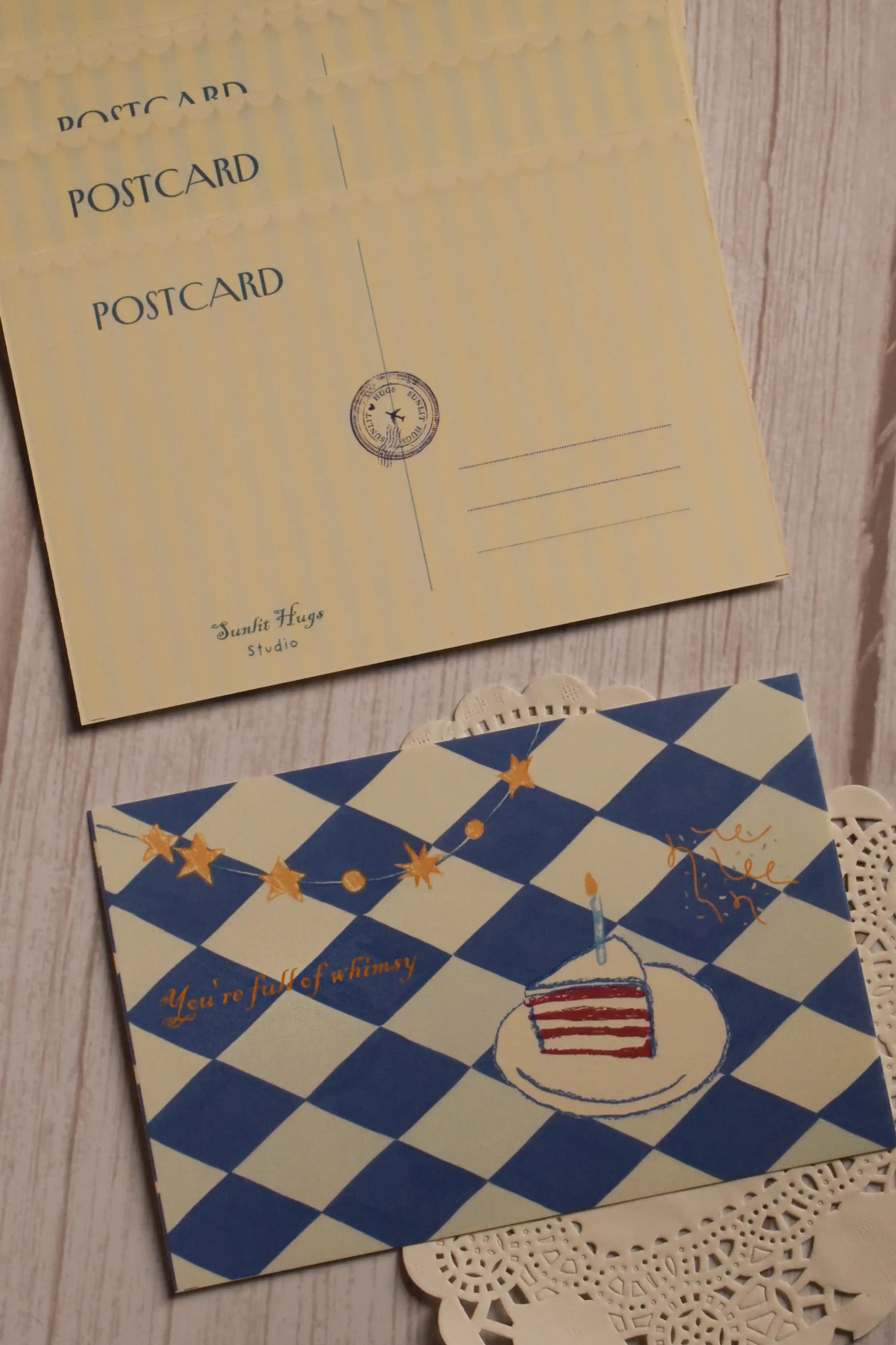 Front and back view of the 4 x 6 inch You’re Full of Whimsy Birthday Postcard featuring a blue and cream harlequin pattern, birthday cake, golden stars and a vintage-inspired writing side.