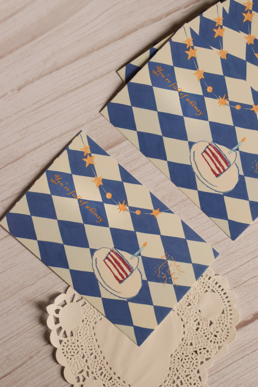 Two whimsical birthday postcards displayed together, showing the blue harlequin pattern, golden stars, confetti and illustrated slice of birthday cake.