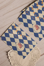 Two whimsical birthday postcards displayed together, showing the blue harlequin pattern, golden stars, confetti and illustrated slice of birthday cake.
