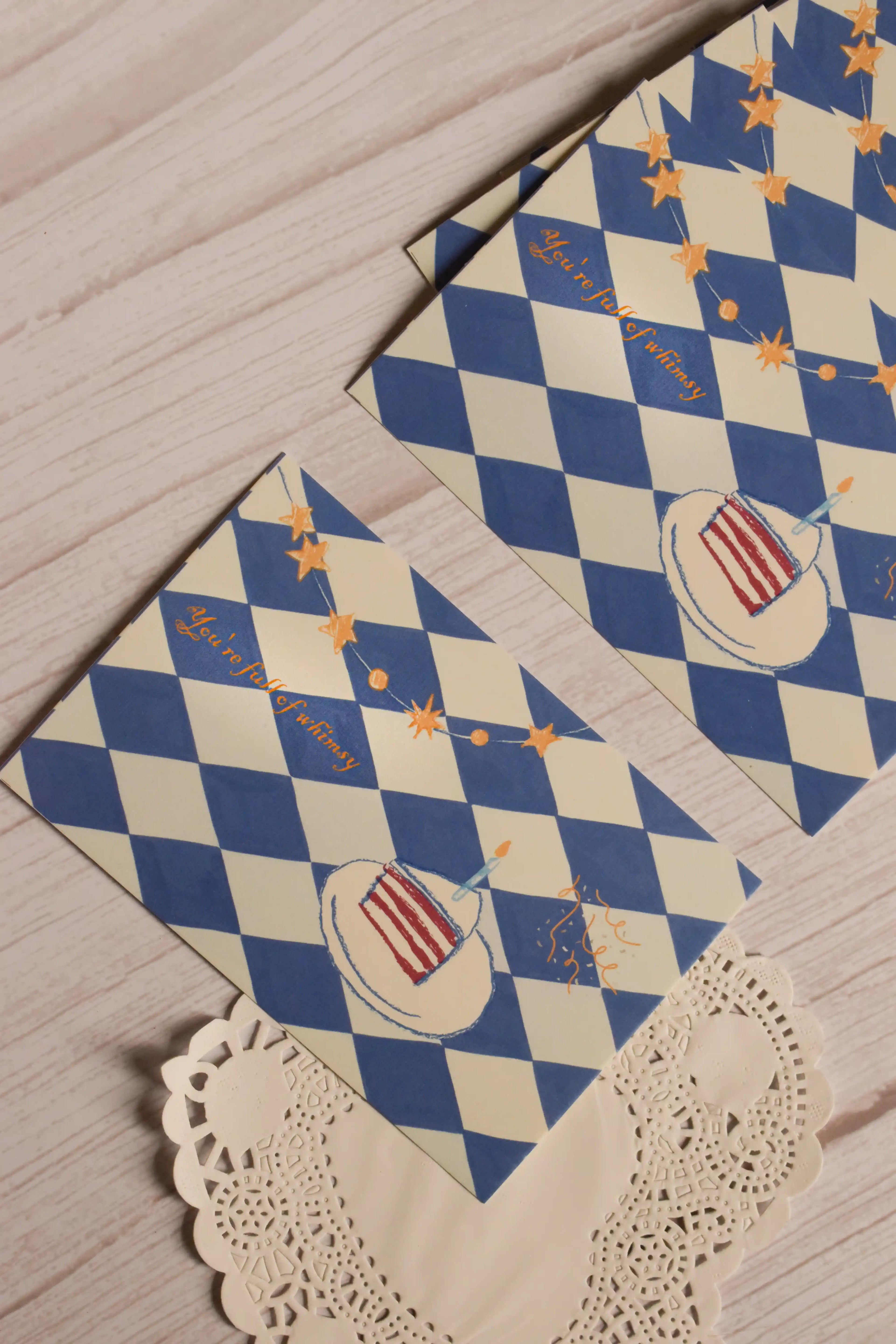 Two whimsical birthday postcards displayed together, showing the blue harlequin pattern, golden stars, confetti and illustrated slice of birthday cake.